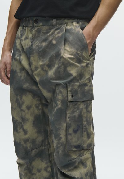 Άνδρας που φοράει μαύρο και πράσινο τσάντι-ντάι παντελόνι cargo με πλαϊνή τσέπη με καπάκι και έχει τα χέρια του μπροστά στις τσέπες.