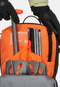 Oranges Innenraum mit einer grauen Schaufel mit Ausschnitten, metallischen Stiften und einem orangefarbenen Notfallsignalwerkzeug, das in einem schwarzen Rucksack aufbewahrt wird.