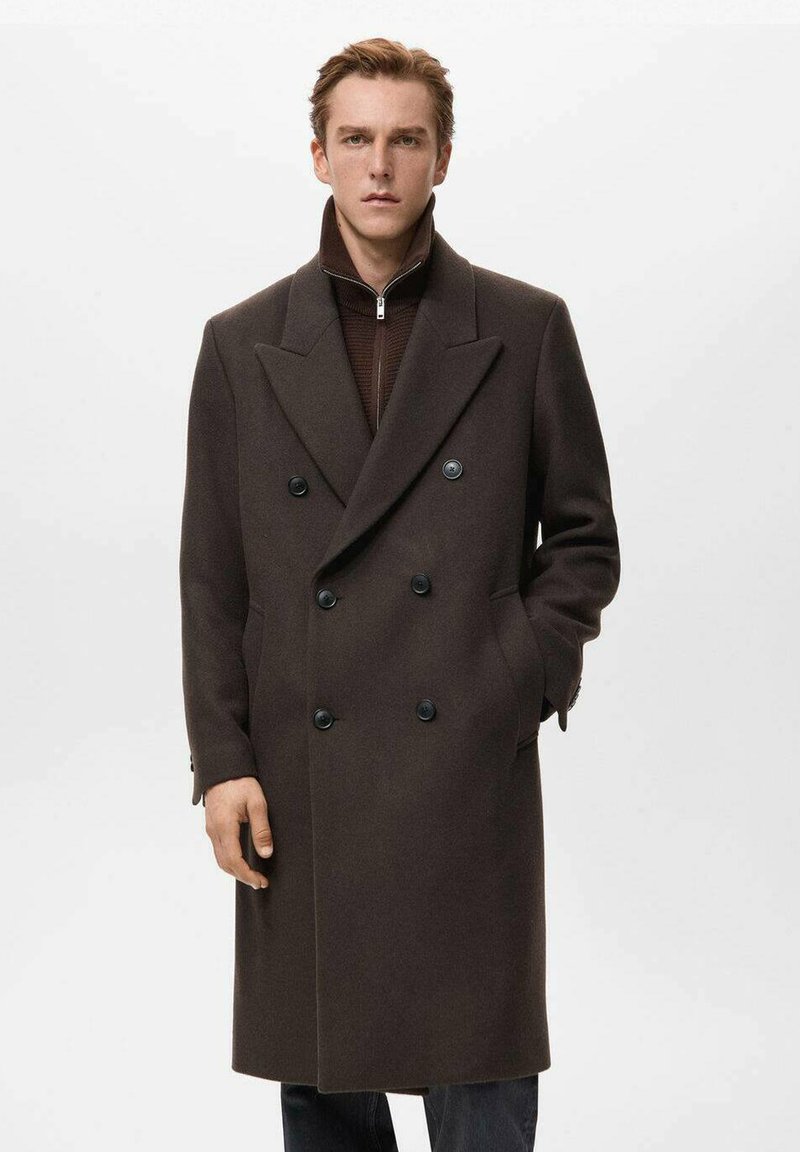 Abrigo marrón de lana con doble botonadura, solapas grandes, cierre de botones y bolsillos laterales. Se lleva sobre un suéter oscuro con cremallera, con una silueta estructurada.