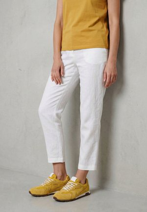 Osoba oblečená v bílých krátkých kalhotách, hořčicovém tílku a hořčicově žlutých teniskách stojí před světle šedou zdí.