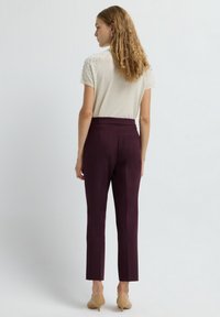 Vrouw met krullend haar in een witte gebreide top met korte mouwen en gestructureerde stippen, hoge pruimenkleurige broek en beige lage hakken, met haar rug naar de kijker gekeerd.