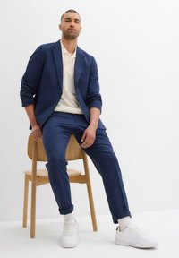 Blazer blu navy e pantaloni abbinati con risvolti, abbinati a una polo beige chiaro e sneakers bianche, seduto su una sedia di legno.