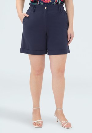 Kobieta ubrana w granatowe szorty z wysokim stanem i podwiniętymi nogawkami, kwiatowy top włożony do środka oraz białe sandały na obcasie z paskami, stoi z rękami w kieszeniach.