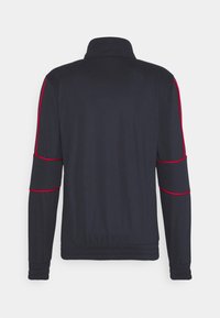 Marinblå jacka med ståkrage, prydd med röd piping längs ärmarna. Elastiska muddar och midja för en figurnära look. Slät textur.