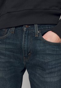 Jeans de mezclilla oscura con una textura sutil, con costuras naranjas, un diseño clásico de cinco bolsillos y una cremallera en la bragueta.
