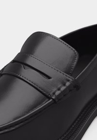 Gros plan sur un mocassin en cuir noir lisse avec des coutures détaillées et une classique bride à monnaie sur la partie supérieure de la chaussure.