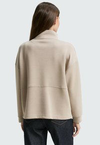 Beige oversized sweatshirt met een hoge kraag, zachte stof en geribbelde manchetten, gecombineerd met donkere denim jeans en zichtbare stikseldetails.