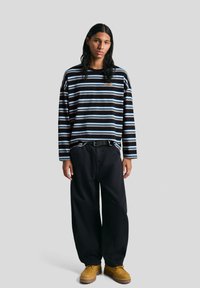 Fekete és világoskék csíkos, hosszú ujjú ing, laza fazonnal, laza fekete nadrággal és sárga lábbeli kiegészítve, egyszerű övvel.