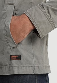 Grå bomuldsjakke med en sidelomme, et Superdry-logo-patch og synlige syninger på ærmerne og kanten.