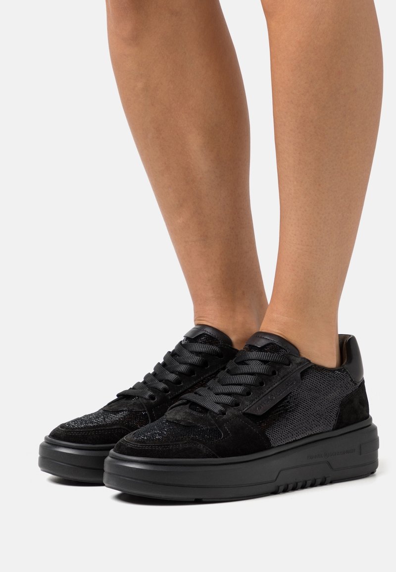Pieds portant des baskets basses noires avec des panneaux texturés et en suède, des semelles épaisses en caoutchouc et des lacets noirs sur un fond clair uni.