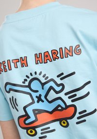 T-shirt in cotone azzurro chiaro con un disegno in outline nero di una figura che fa skateboard, con accenti rossi e arancioni, e il testo "KEITH HARING."