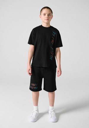 Junge steht und trägt ein schwarzes T-Shirt und Shorts mit buntem "Paul Smith"-Schriftzug, weiße Socken und weiße Turnschuhe vor hellem Hintergrund.