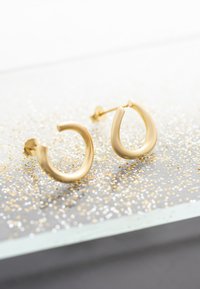 Guldörhängen i form av hoops med en slät, rundad design. De har ett stiftbaksidan och en spegelbeläggning på en glittrig bakgrund.