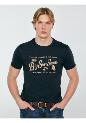 Młody mężczyzna z blond włosami w czarnej koszulce "Big Star Jeans" i niebieskich dżinsach, z rękami w kieszeniach, na tle jasnej, jednolitej ściany.