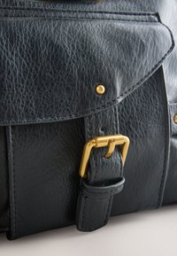 Melna ādas soma tuvplānā ar teksturētu virsmu, zelta toņa sprādzi un šūtu pārsegu. Komplektā ar kniedes detaļām.