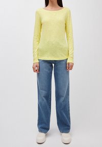 Person, der ein hellgelbes Langarmshirt, blaue weite Jeans und weiße Slipper trägt, steht vor einem schlichten Hintergrund.