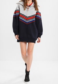Mujer vistiendo un vestido sudadera largo y oversized a rayas en navy, gris, rojo, blanco y azul, con zapatillas negras de caña alta, de pie con una pierna cruzada.