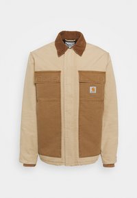 Beige en bruine Carhartt-jack met een corduroy kraag, twee grote voorkruiken en een zichtbaar logo-label op de rechter zak.