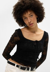 Μαύρη μακρυμάνικη μπλούζα από διάφανο λουλούδι με τετράγωνη λαιμόκοψη και κορδόνι μπροστά. Συνδυάζεται με μια μαύρη ζώνη με ασημένια υλικά.