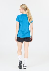 Kvinna med långt blont hästsvans iförd en blå skjorta, svarta shorts, vita strumpor och sneakers, ståendes vänd bort från kameran mot en vit bakgrund.