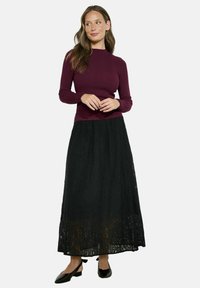 Sieviete, tērpusies ribotas struktūras, garām piedurknēm bordo krāsas blūzē, melnā mežģīņu A-formas garā svārkā un melnās siksnu zema papēža kurpēs, stāv ar rokām saliktām kopā.