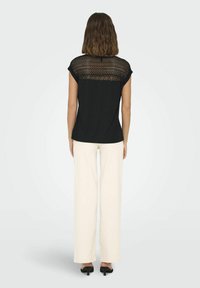 Czarna koszula z krótkim rękawem i koronkowym detalem na plecach, zestawiona z kremowymi spodniami typu szerokie nogawki. Gładka tekstura i luźny fason.