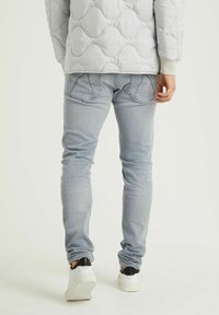 Person som bär ljusgrå jeans och en vadderad ljusgrå jacka, vita sneakers med svart häl, står på en ljus bakgrund, bakifrån.