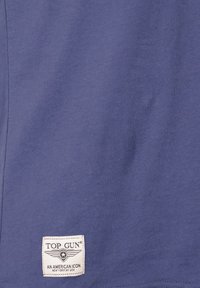 Lilla bomulds T-shirt med et hvidt mærke med "TOP GUN" og et emblem med vinger. Stoffet ser glat og let ud.