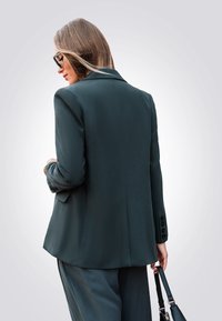 Donkergroene op maat gemaakte blazer met een enkelvoudige sluiting, slim fit en zwarte knopen; heeft een gladde textuur en een split aan de achterkant.