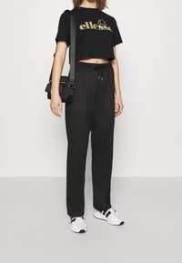 Černé zkrácené tričko s zlatým logem, černé volné kalhoty a bílé tenisky s černými akcenty. Malá černá crossbody taška.