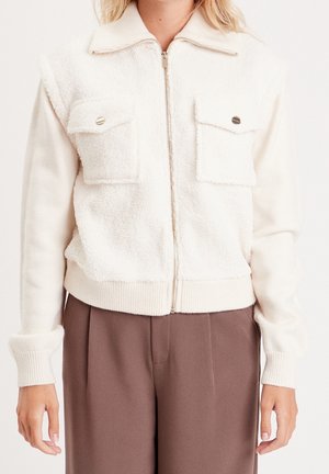 Femme portant une veste polaire blanche zippée avec deux poches poitrine et un pantalon marron taille haute, vue du cou aux hanches.