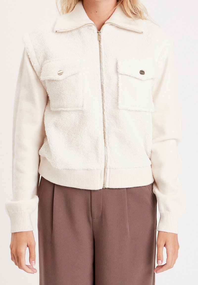 Femme portant une veste polaire blanche zippée avec deux poches poitrine et un pantalon marron taille haute, vue du cou aux hanches.