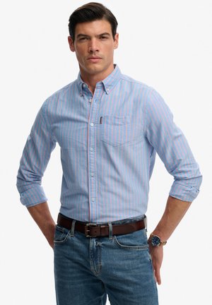 Mężczyzna w jasnoniebieskiej koszuli w różowe paski, niebieskich dżinsach, brązowym pasku i zegarku na ręku, stojący z rękami za plecami.