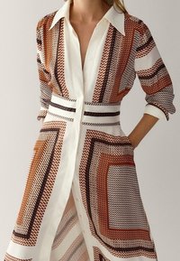 Robe chemise longue avec une base blanche et des motifs géométriques en rouille, marron et noir. Fini avec une fermeture à boutons et un col.