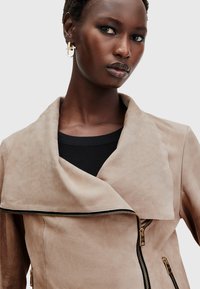 Giacca in suede beige con colletto drappeggiato, zip frontale asimmetrica e dettagli in hardware dorato. Top nero a coste sotto.