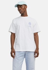 Camiseta blanca de algodón con mangas cortas, que presenta un logo y diseño en azul claro en el lado izquierdo del pecho. El modelo lleva una gorra azul clara y jeans de mezclilla.