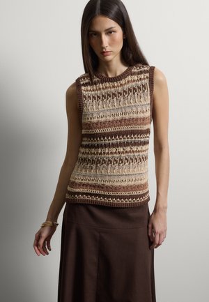 Jeune femme aux cheveux longs et foncés portant un haut sans manches en tricot avec des motifs marron, beige et crème, ainsi qu'une jupe marron foncé.