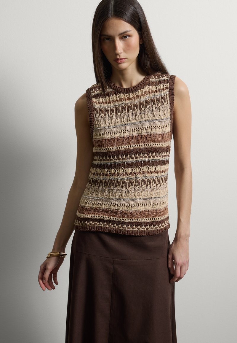 Junge Frau mit langen dunklen Haaren, die ein ärmelloses, gestricktes Oberteil mit braunen, beigen und cremefarbenen Mustern sowie einen dunkelbraunen Rock trägt.