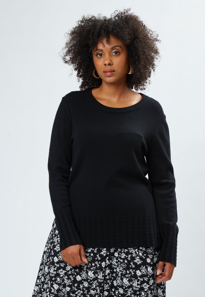 Sort strikket sweater med rund hals og ribbede manchetter, parret med en sort og hvid blomstret nederdel. Enkel, struktureret design.
