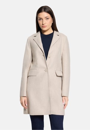 Kvinde med lige mørkt hår iført en beige knapjakke over en sort top og mørke bukser, stående mod en ensfarvet baggrund.