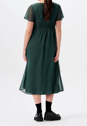 Femme vue de dos portant une robe midi vert foncé avec des manches courtes transparentes et des mocassins noirs avec des chaussettes noires.
