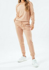 Béžová mikina a odpovídající tepláky vyrobené z měkké tkaniny, s logem vpředu. Nosit s bílými teniskami. Jednoduchý, tapered střih.