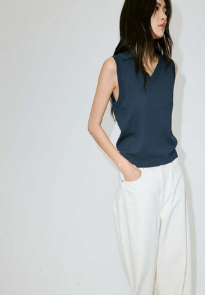 Femme aux longs cheveux foncés portant un top sans manches bleu marine et un pantalon blanc à jambes larges, debout avec une main dans la poche.