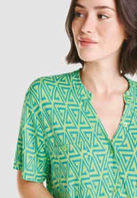 Blusa verde de manga corta con un patrón geométrico azul, escote en V y un único bolsillo en el pecho con un detalle de botón a juego.