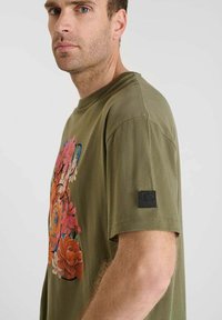 Camiseta verde oliva con un vibrante diseño gráfico floral, mangas cortas y un parche de acento negro en la manga izquierda.