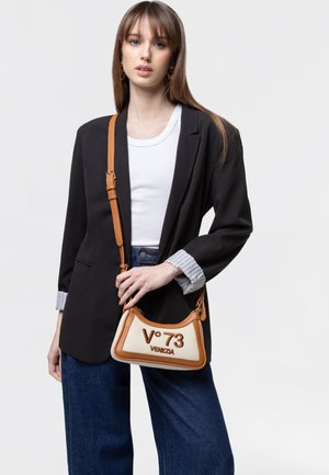 Jonge vrouw draagt een zwarte blazer, wit topje en blauwe jeans, houdt een beige en bruine handtas vast met "V°73 Venezia" aan de voorkant.