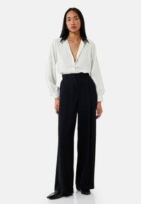 Blusa blanca con cuello negro, abotonada, combinada con pantalones negros de pierna ancha y cintura alta, acompañada de zapatos negros brillantes.