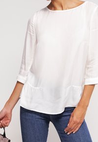 Blusa bianca leggera con scollatura a barca, maniche a tre quarti e dettaglio in vita arricciato. Indossata con jeans blu.