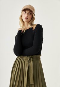 Suéter negro con canalé y botones en el hombro, combinado con una falda verde olivo plisada que presenta una cintura atada y una cinturilla decorativa.