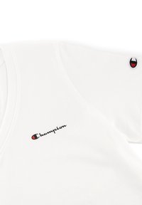 T-shirt bianco in cotone con scollatura a V. Presenta un logo nero "Champion" e un piccolo emblema ricamato rosso e nero sulla manica.